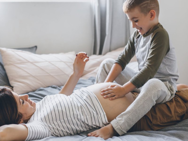 Schwangere Frau liegt auf dem Bett, ihr älterer Sohn, ca. 5 Jahre alt, sitzt auf ihrem Schoß und streichelt den Bauch.