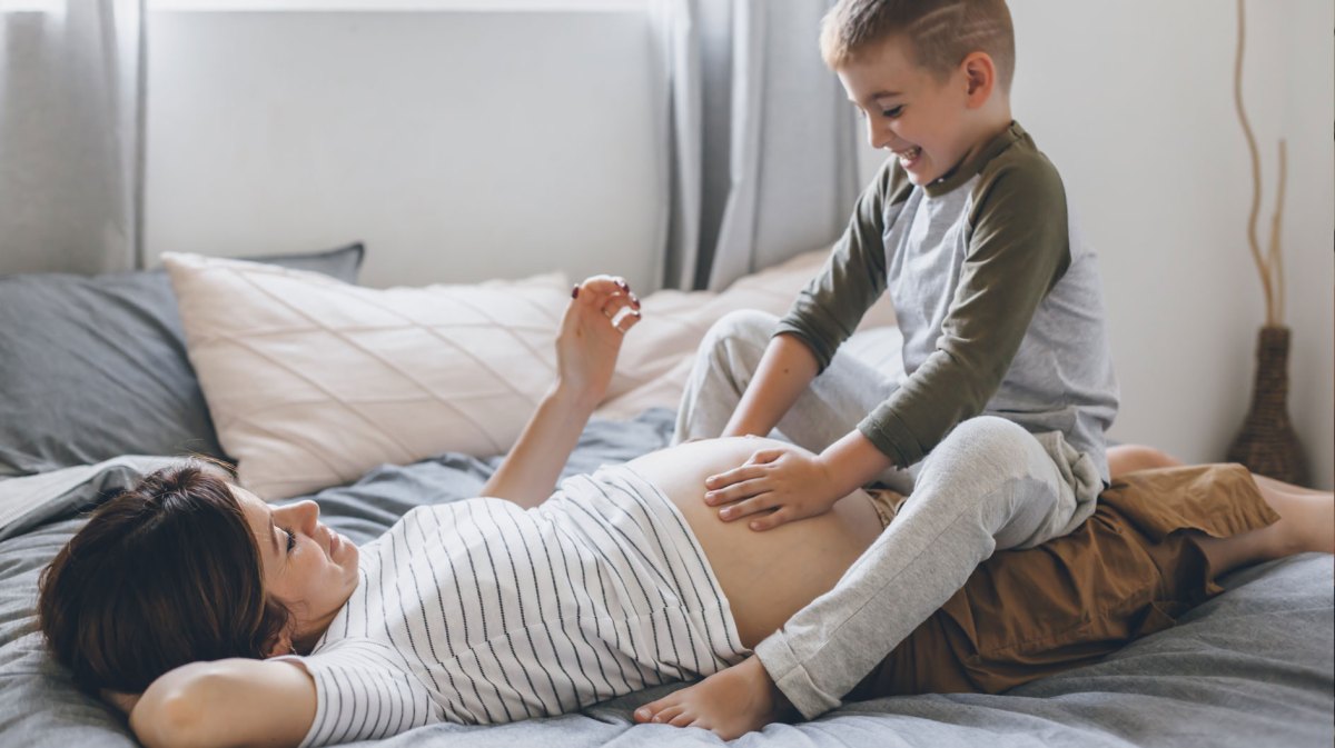 Schwangere Frau liegt auf dem Bett, ihr älterer Sohn, ca. 5 Jahre alt, sitzt auf ihrem Schoß und streichelt den Bauch.