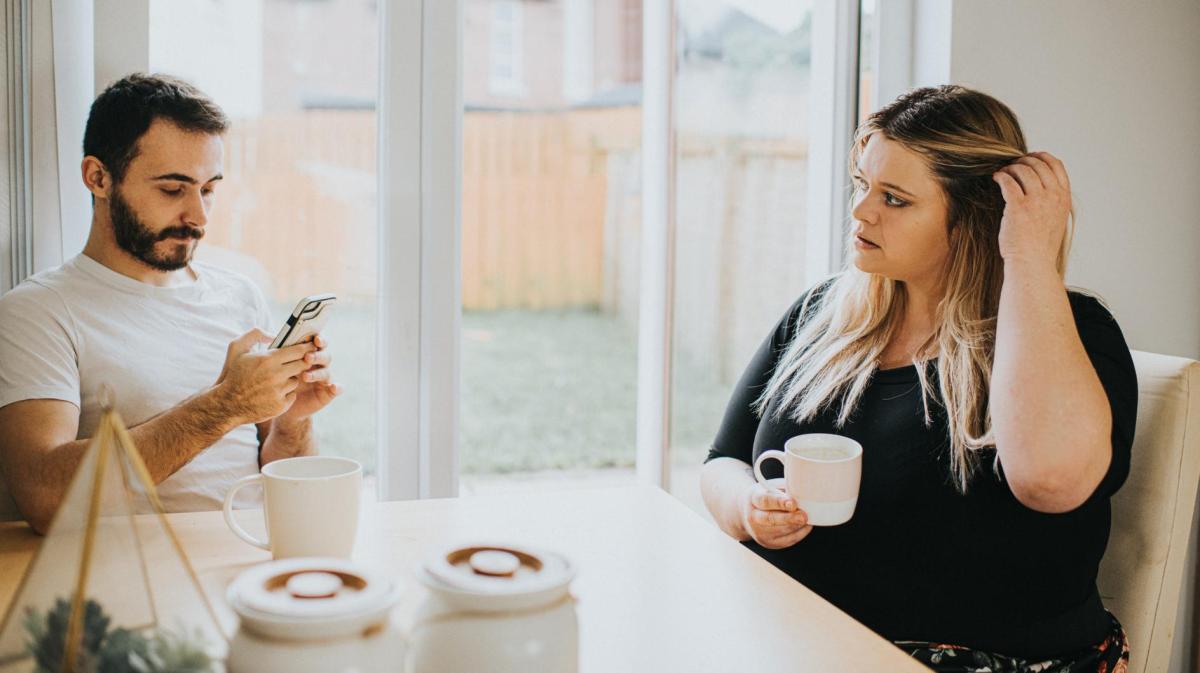 Ein Mann sitzt am Handy und seine Frau sitzt mit einer Tasse gegenüber und guckt ihn an.