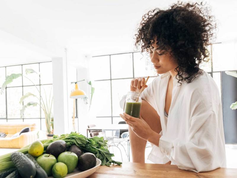 Junge hübsche Frau entspannt sich in der Küche, während sie einen grünen Saft mit Bambusstrohhalm am Morgen trinkt. Entgiften. Reinigen. Fasten. Detox.