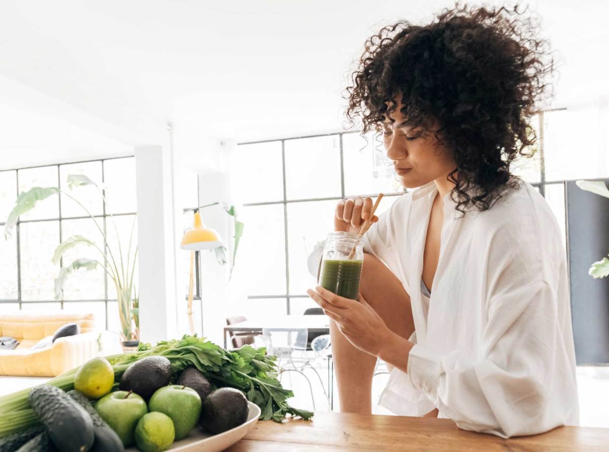 Junge hübsche Frau entspannt sich in der Küche, während sie einen grünen Saft mit Bambusstrohhalm am Morgen trinkt. Entgiften. Reinigen. Fasten. Detox.