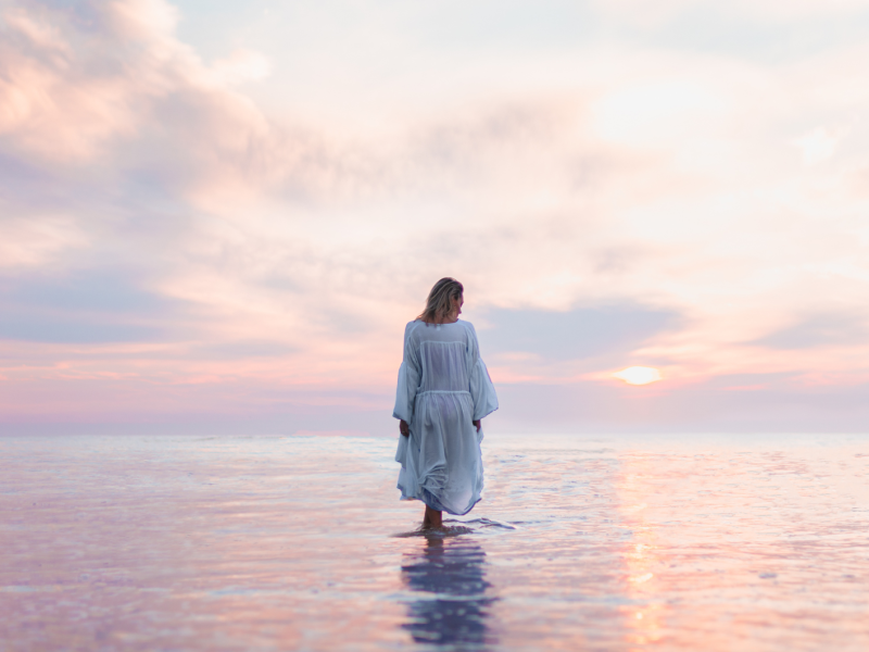 Frau bei Sonnenuntergang, die ein weißes Kleid anhat und auf Wasser geht.