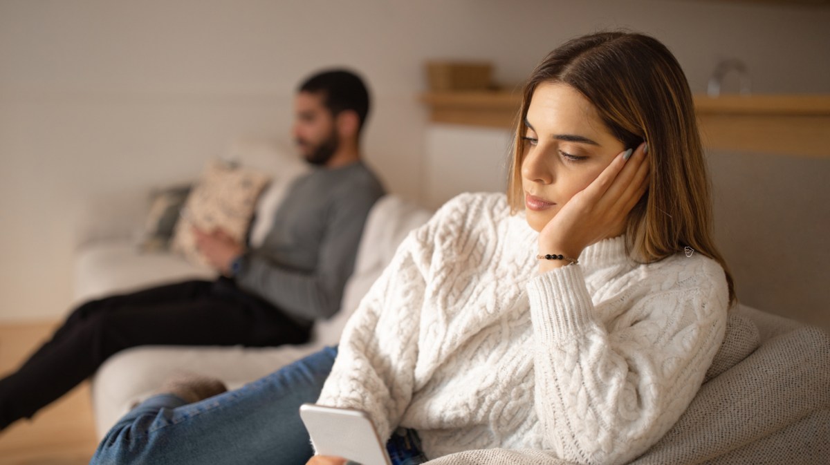 Frau auf einem Sofa, die bedrückt nach unten auf ihr Handy schaut, während ein Mann im Hintergrund sitzt.