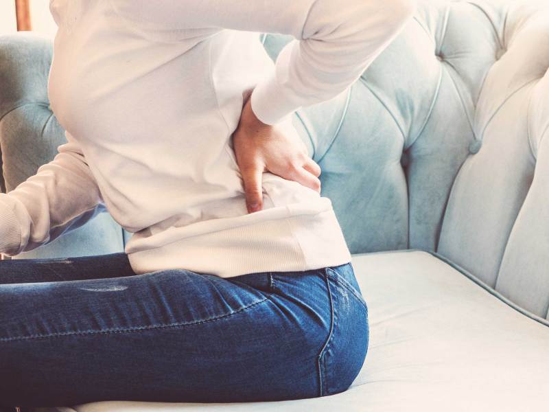 Weibliche Hände berühren schmerzenden unteren Rücken und Po, sitzend auf Sofa zu Hause.