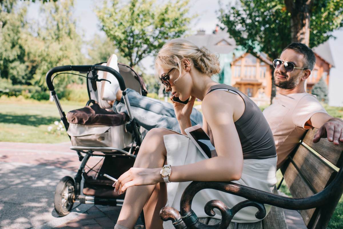 Paar sitzt auf einer Parkbank, er hat die Beine lässig auf dem Kinderwagen abgelegt, sie sitzt neben ihm, raucht und telefoniert am Handy.