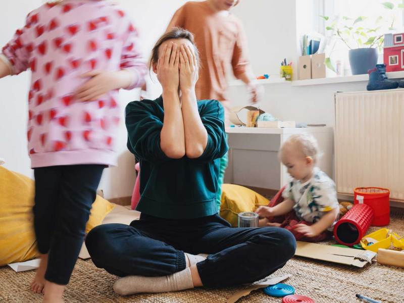 Mutter sitzt im Schneidersitz und mit Händen vor das Gesicht geschlagen auf dem Boden, um sie herum spielen drei Kinder unterschiedlichen Alters.