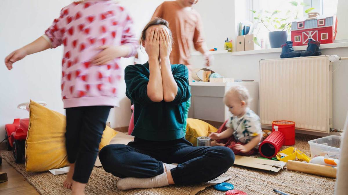 Mutter sitzt im Schneidersitz und mit Händen vor das Gesicht geschlagen auf dem Boden, um sie herum spielen drei Kinder unterschiedlichen Alters.