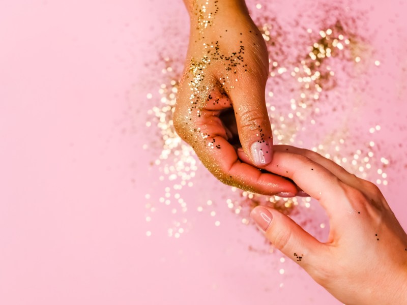 Zwei Hände mit GLitzer vor rosa Hintergrund, die sich an den Fingerspitzen berühren und anfassen