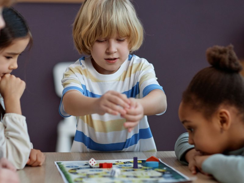 4 Kinder der ersten Klasse sitzen an einem Tisch und spielen ein Würfelspiel.