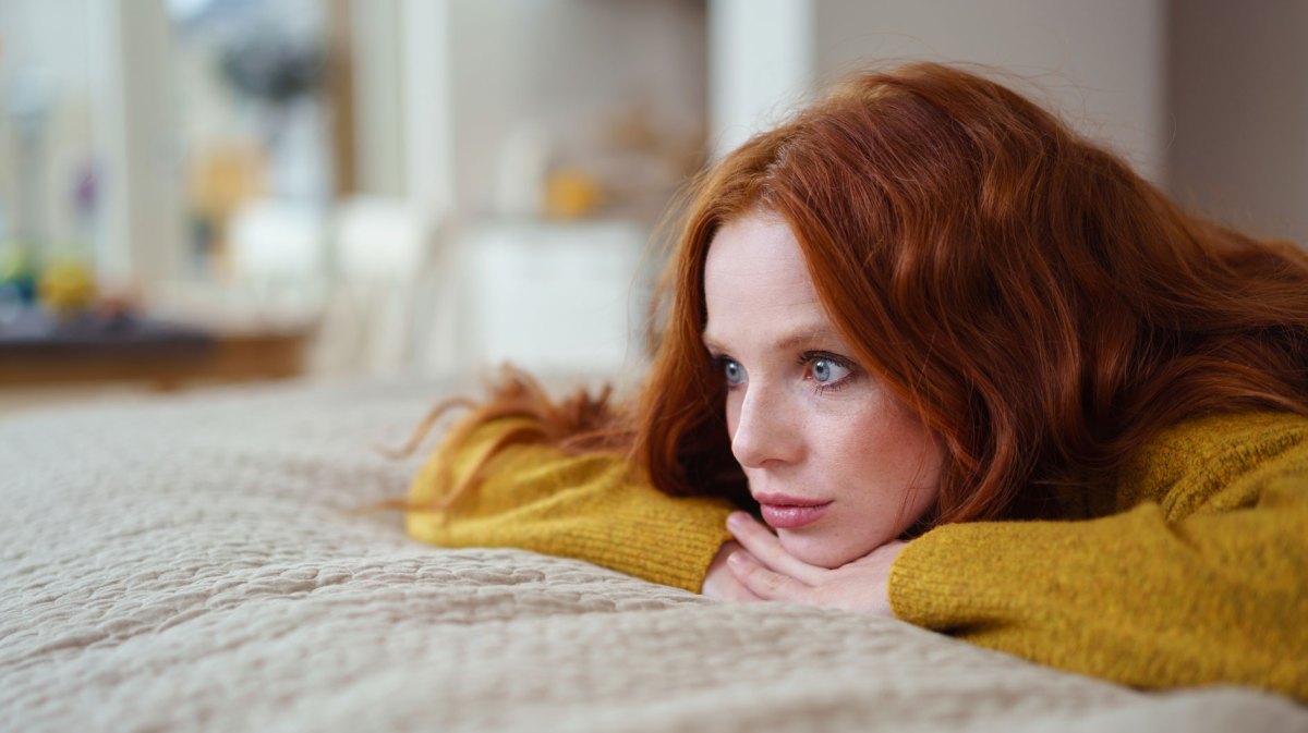 Frau mit roten Haaren und gelbem Pullover liegt auf dem Bett und blickt ernst.