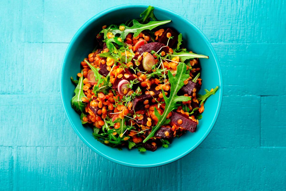 Türkise Schale mit gesundem Salat aus Linsen, Rote Beete und Rucola, angerichtet auf blauem Holztisch.