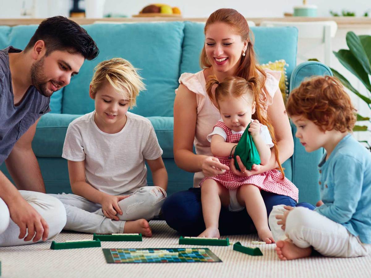 Mutter, Vater und die drei Kinder unterschiedlichen Alters spielen eine Runde Scrabble auf dem Fußboden.