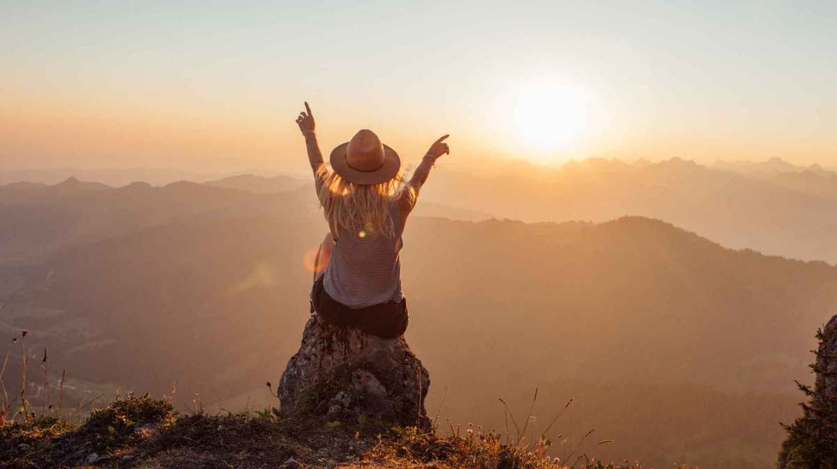 Frau mit Hut sitzt auf einem Felsen vor einer Landschaft.