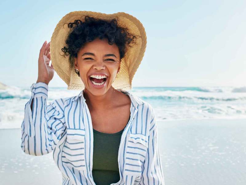 Junge Frau mit dunklen Haaren und Sonnenhut steht am Strand und lacht fröhlich.