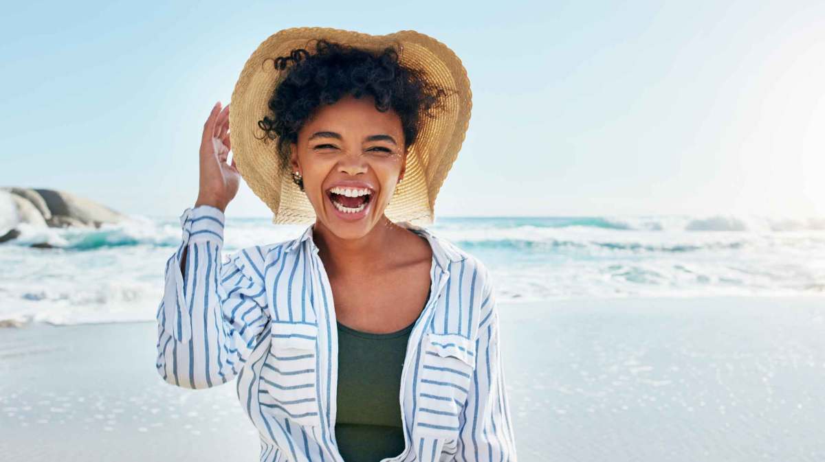 Junge Frau mit dunklen Haaren und Sonnenhut steht am Strand und lacht fröhlich.