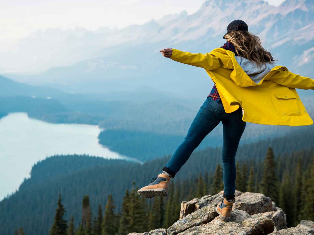 Frau steht auf einem Fels vor einer Seen- und Berg-Landschaft.