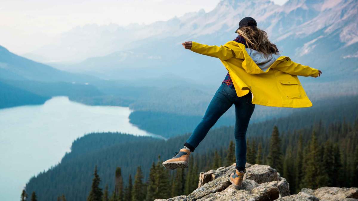 Frau steht auf einem Fels vor einer Seen- und Berg-Landschaft.