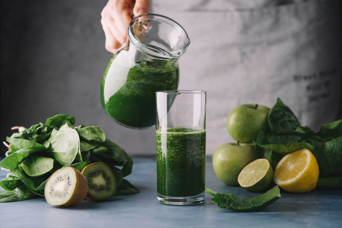 Weibliche Hände gießen grünen Smoothie in ein Glas. Zutaten: Spinat, Apfel, Kiwi, Zitrone, Limette. Detox-Frühstück