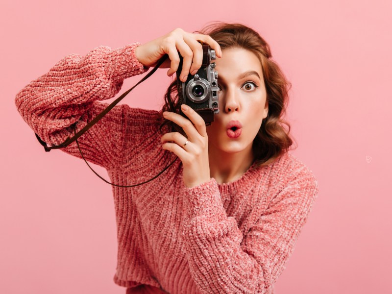 Frau vor pinkem Hintergrund, die eine Kamera in der Hand hält und überrascht guckt.
