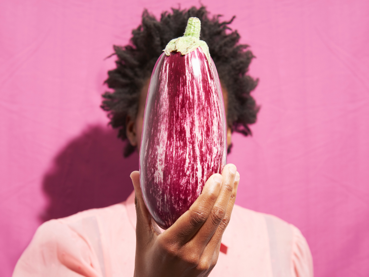 Eine gestreifte Aubergine wird von einer Person mit Afrofrisur vor einem knallpinken Hintergrund hochgehalten.