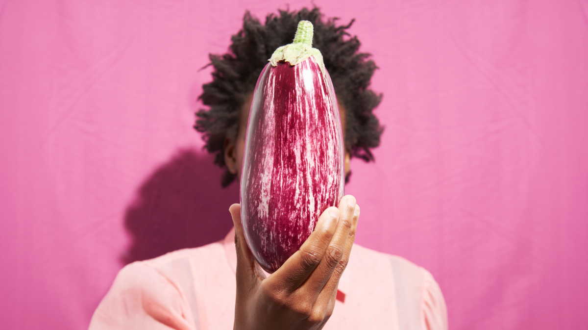 Eine gestreifte Aubergine wird von einer Person mit Afrofrisur vor einem knallpinken Hintergrund hochgehalten.