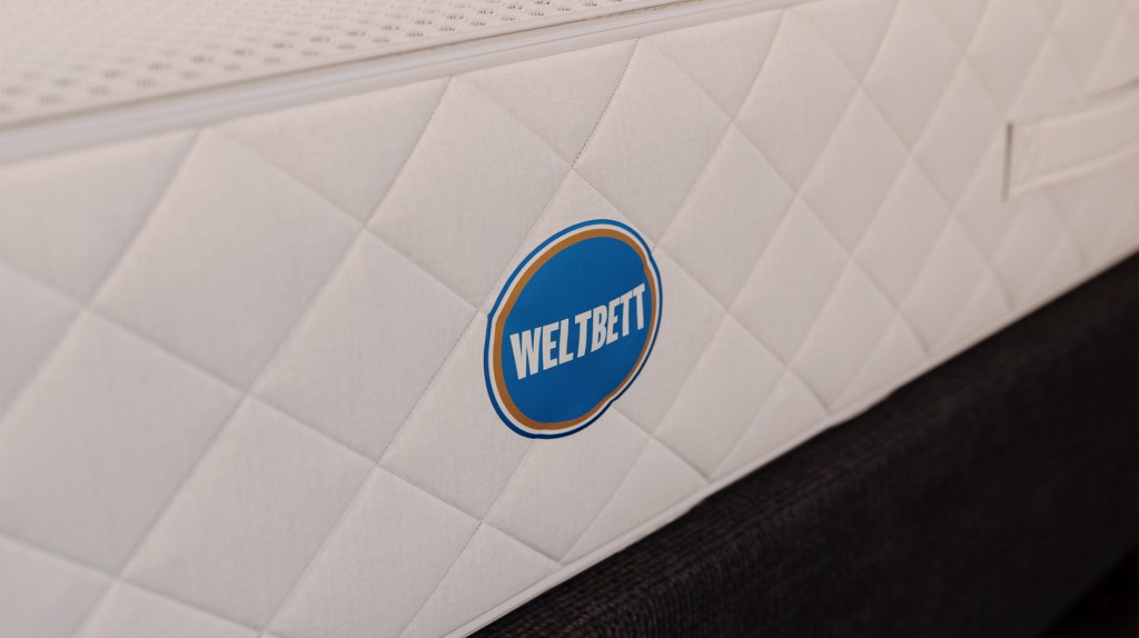 Detailaufnahme einer weißen Matratze mit gestepptem Bezug und „Weltbett“-Logo, platziert auf einem dunklen Stoffbettgestell.