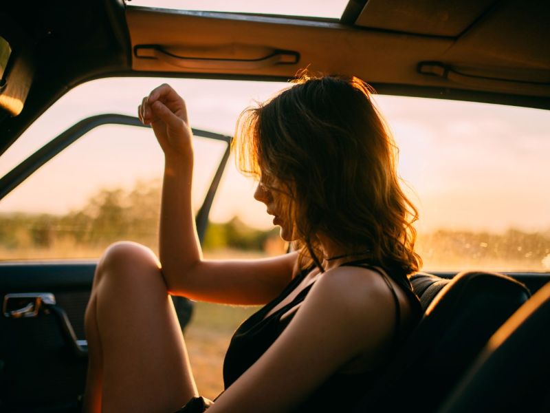 Junge Frau mit dunklen Haaren sitzt bei Sonnenuntergang im Auto bei offener Tür.