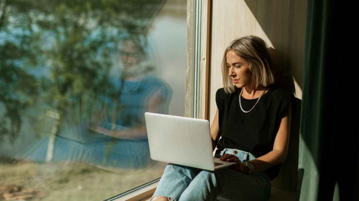 Frau sitzt am Fenster vor ihrem Laptop.