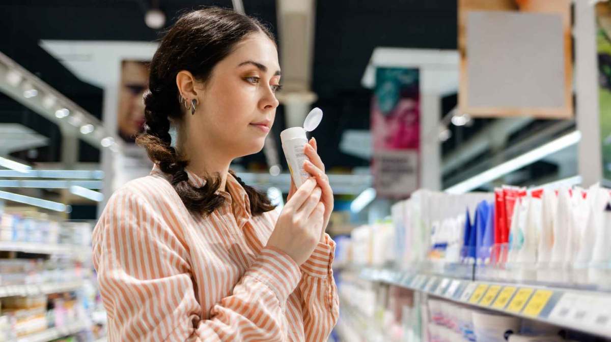 Junge Frau mit dunklen Haaren steht im Supermarkt und riecht an einem Shampoo.