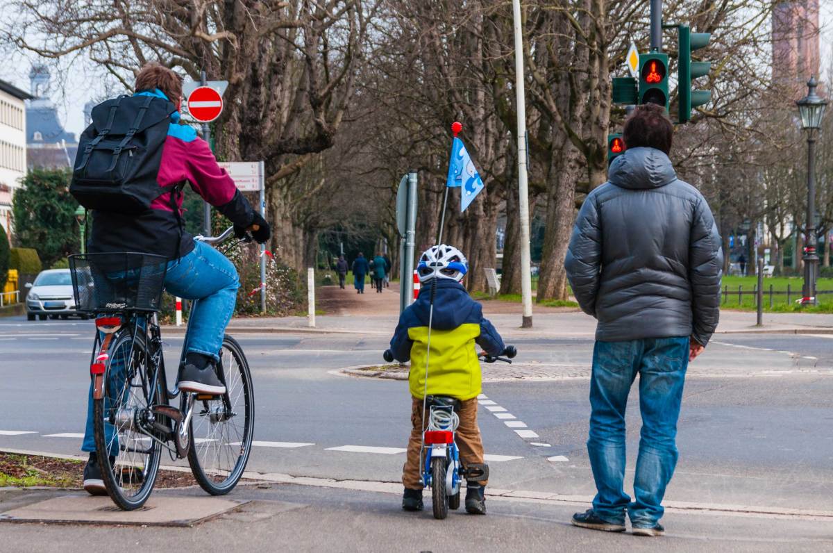 Vater und ca. 4-jähriges Kind stehen auf ihren Fahrrädern sitzend an der Ampel und warten auf Grün.