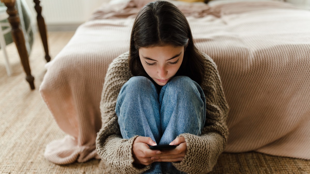 Frau, die vor ihrem Bett sitzt und schüchtern auf ihr Handy starrt