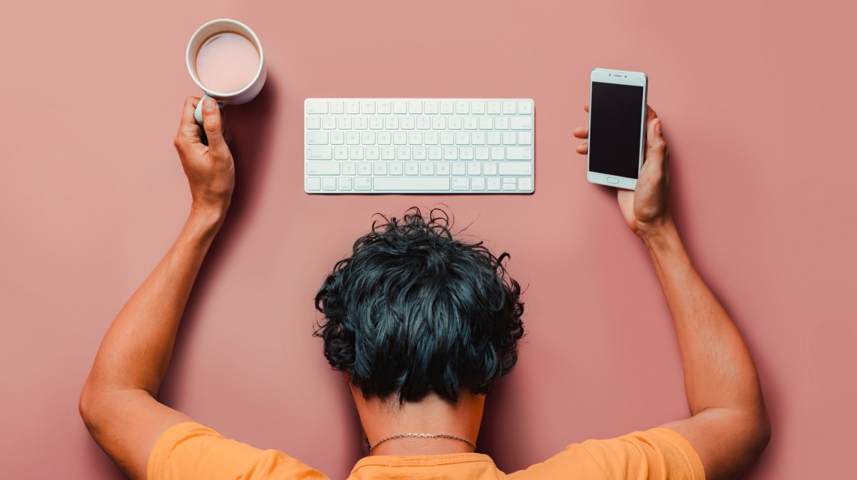 Mann vor rosa Hintergrund, der ein Handy und einen Kaffee in der Hand hält, während sein Kopf vor der Tastatur auf dem Boden liegt