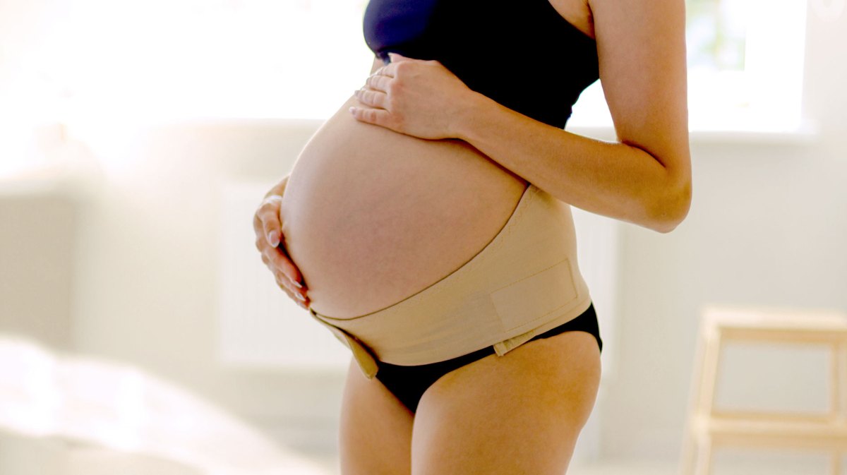 Hochschwangere Frau hat sich einen breiten Klettgürtel um das Becken gelegt, welcher den großen Babybauch stützt.