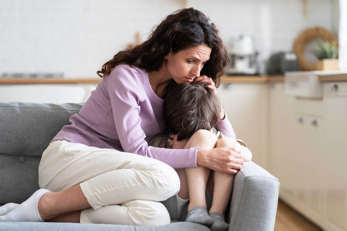 Mutter und Sohn sitzen auf dem Sofa. Er versteckt seinen Kopf zwischen seinen Beinen und seine Mutter tröstet ihn und hält ihn fest im Arm.