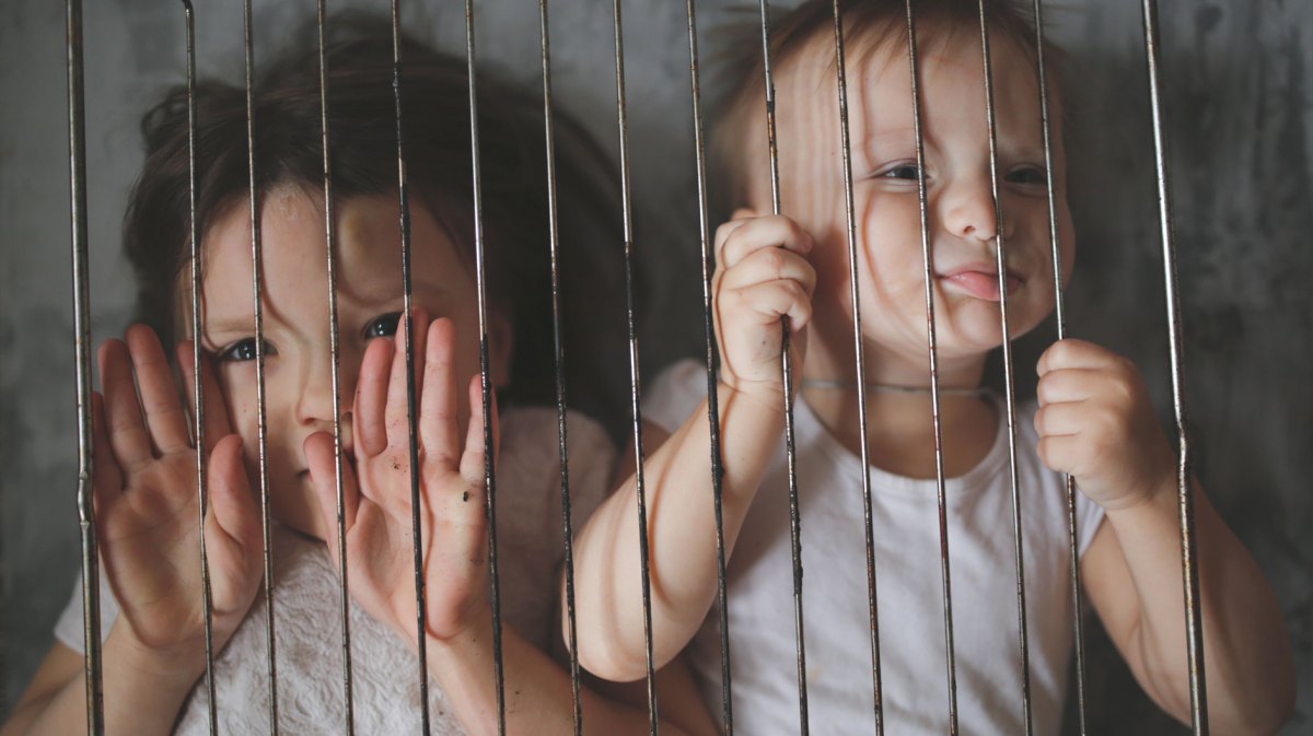 Zwei Kinder, ca. 4 und 2 Jahre alt, liegen auf dem Bett, vor ihren Gesichtern halten sie ein Ofengitter.