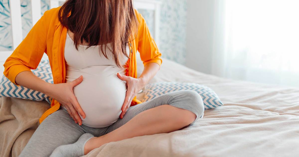 Hochschwangere Frau sitzt auf dem Bett und hält ihren Bauch an den Seiten mit beiden Händen fest, als würde sie einen leichten Schmerz verspüren.