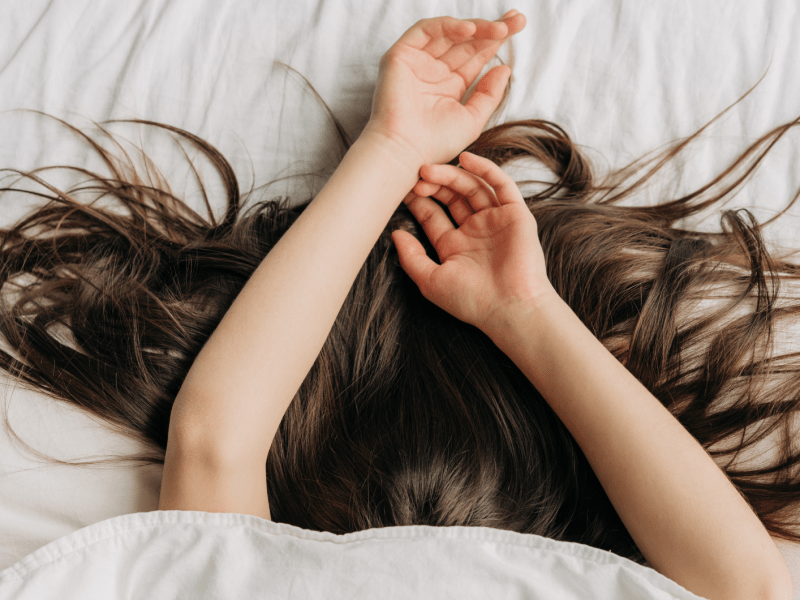 Frau im Bett, die sich unter der Decke versteckt und mit den Armen und dunkelbraunen Haaren herausguckt.