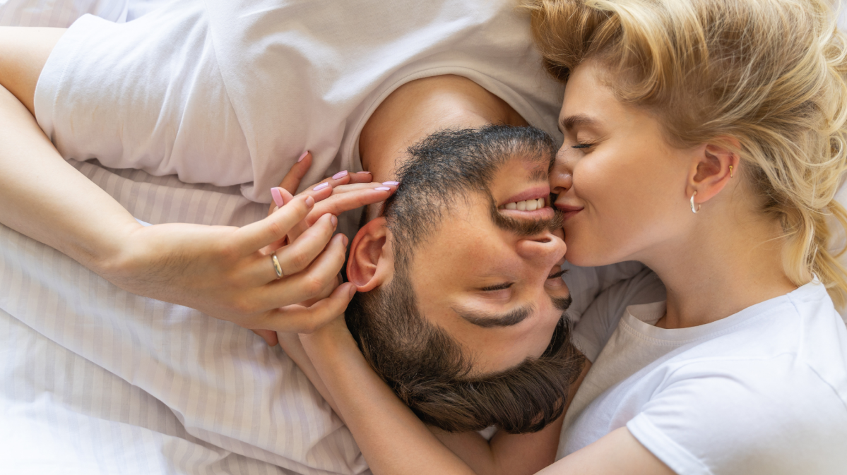 Frau neben Mann im Bett, die ihn auf die Wange küsst.