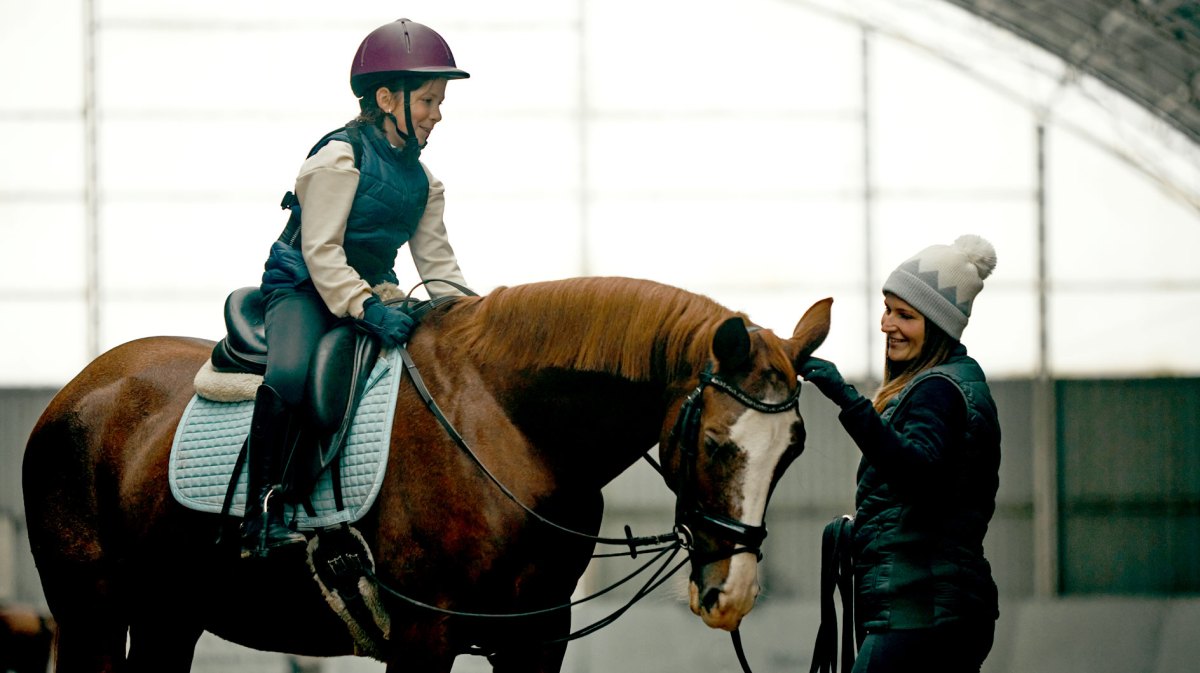 Mädchen auf einem Pferd in einer Reithalle. Die Reitlehrerin steht neben dem Pferd und streichelt es am Kopf.