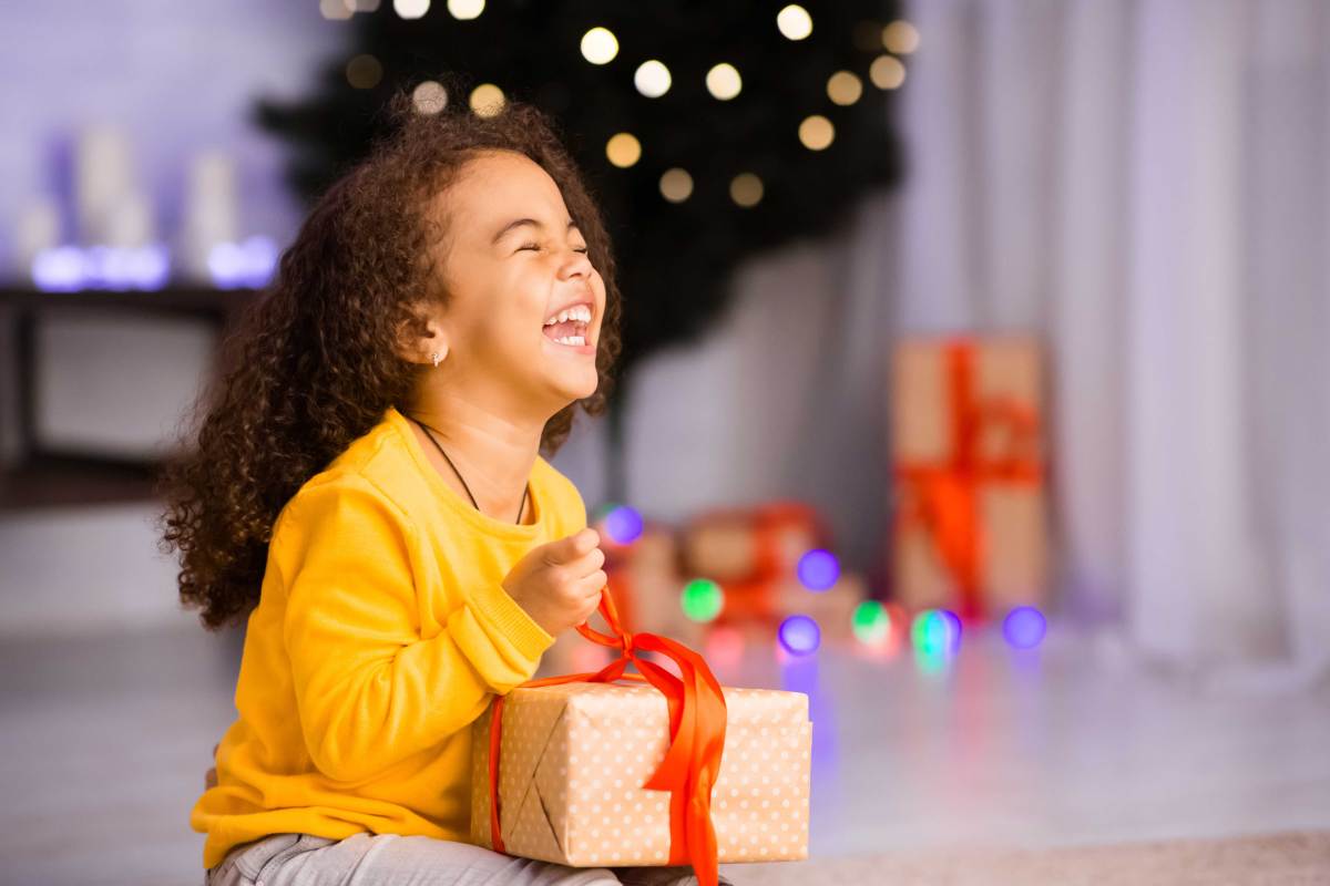 Kleines Mädchen sitzt vorm Weihnachtsbaum und packt sehr aufgeregt ein Geschenk aus.