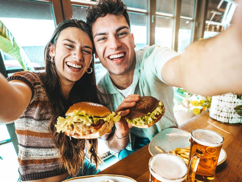 Junges Paar im Restaurant nimmt ein Selfie auf. Beide halten einen Burger in der Hand und haben Bier und Pommes vor sich auf dem Tisch stehen.