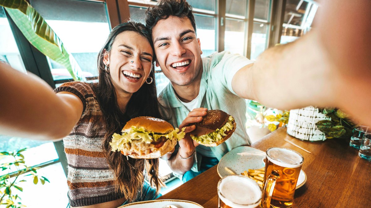 Junges Paar im Restaurant nimmt ein Selfie auf. Beide halten einen Burger in der Hand und haben Bier und Pommes vor sich auf dem Tisch stehen.