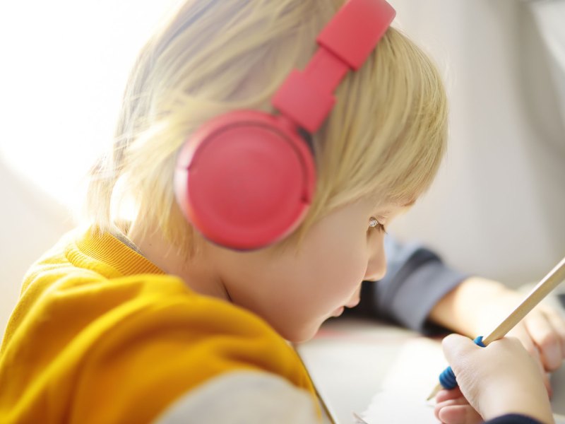 Kind, ca. 5 Jahre, sitzt im Flugzeug, hat Kopfhörer auf und zeichnet gedankenversunken auf einem Blatt Papier.