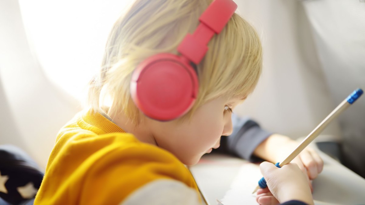 Kind, ca. 5 Jahre, sitzt im Flugzeug, hat Kopfhörer auf und zeichnet gedankenversunken auf einem Blatt Papier.