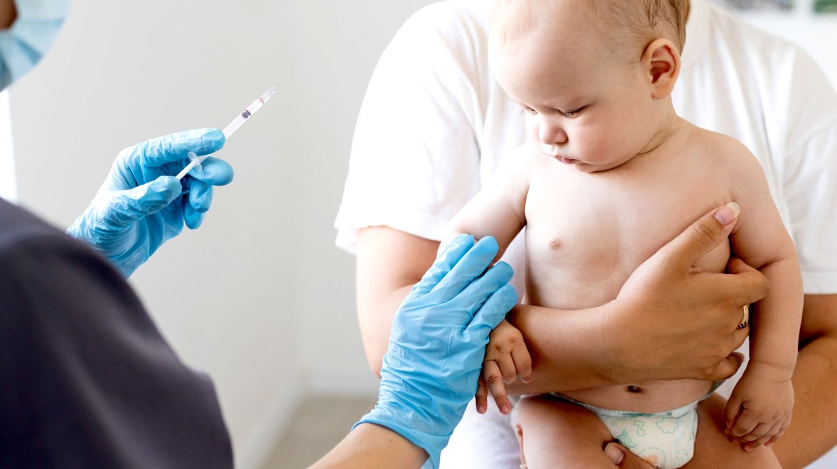 Kleines Baby hat gerade eine Impfung bekommen und schaut skeptisch zur Einstichstelle. Es wird von seinem Vater gehalten.