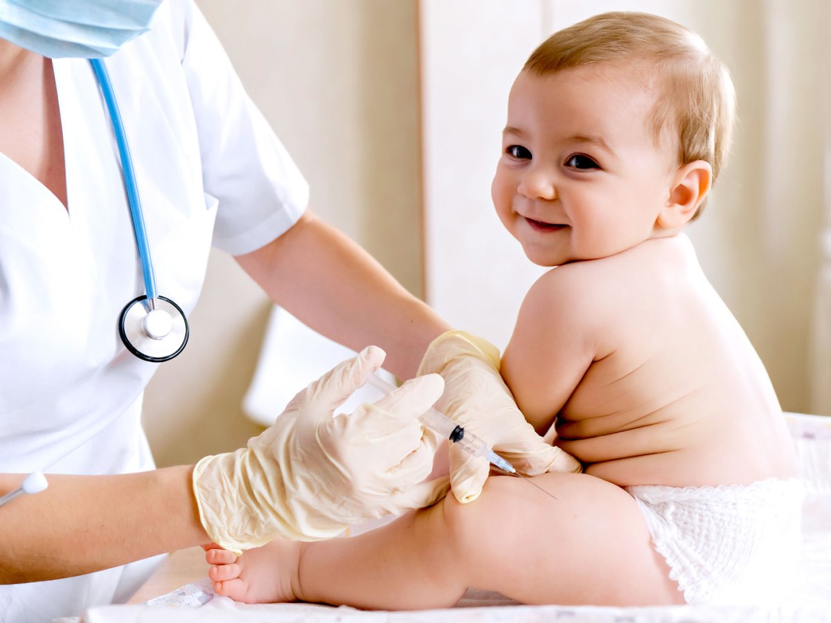 Baby sitzt selbstständig auf der Liege in einem Behandlungsraum und erhält eine Impfung in den Oberschenkel, lacht dabei aber fröhlich in die Kamera.