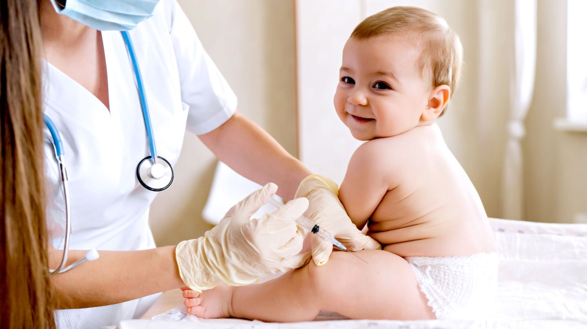 Baby sitzt selbstständig auf der Liege in einem Behandlungsraum und erhält eine Impfung in den Oberschenkel, lacht dabei aber fröhlich in die Kamera.