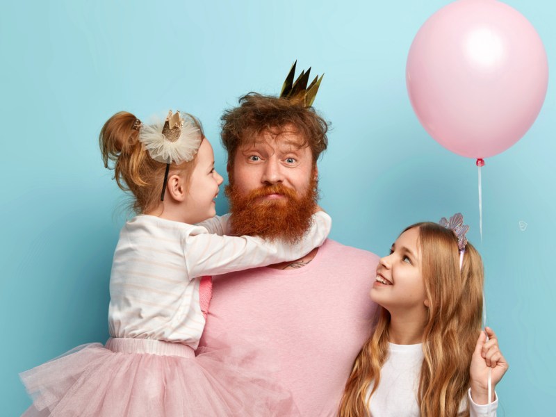 Vater mit zwei Kindern vor einer blauen Wand, der eine Krone aufhat und einen Ballon in der Hand hält.