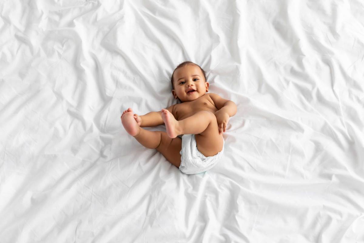 Foto eines Babys von oben, das fröhlich auf einer Decke liegt und mit seinen Füßen spielt.