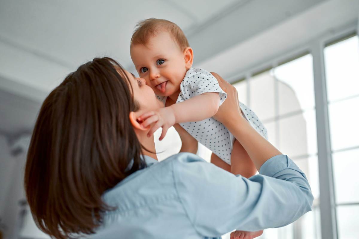 Mutter hebt ihr Baby nach oben und das streckt freudig die Zunge raus.